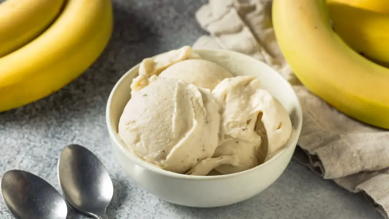 Un cremoso helado de plátano servido en un cuenco blanco, con plátanos amarillos frescos al fondo.