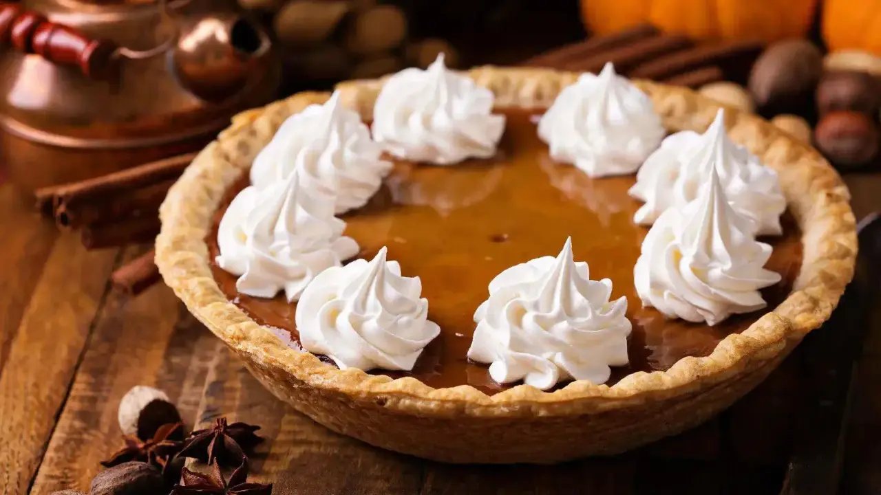 Pastel de calabaza decorado con crema batida, listo para los postres de Acción de Gracias. Especias y calabazas al fondo.