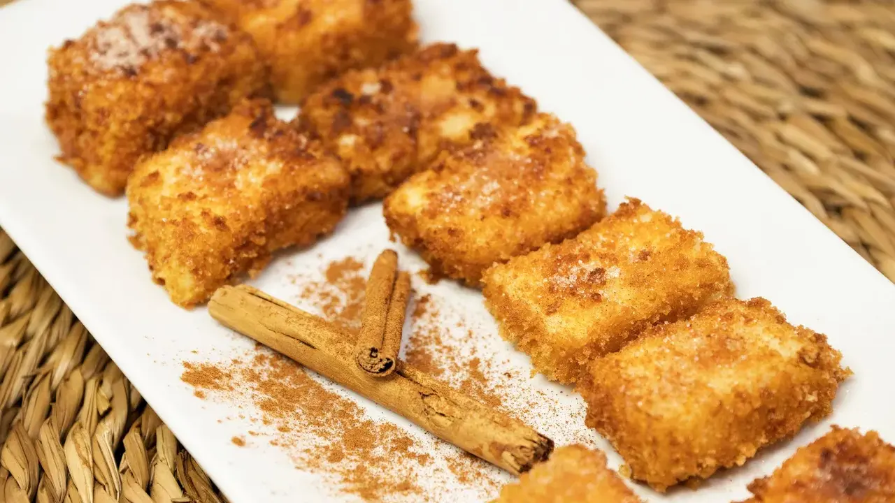Deliciosa leche frita dorada y crujiente, espolvoreada con azúcar y canela, junto a una rama de canela.