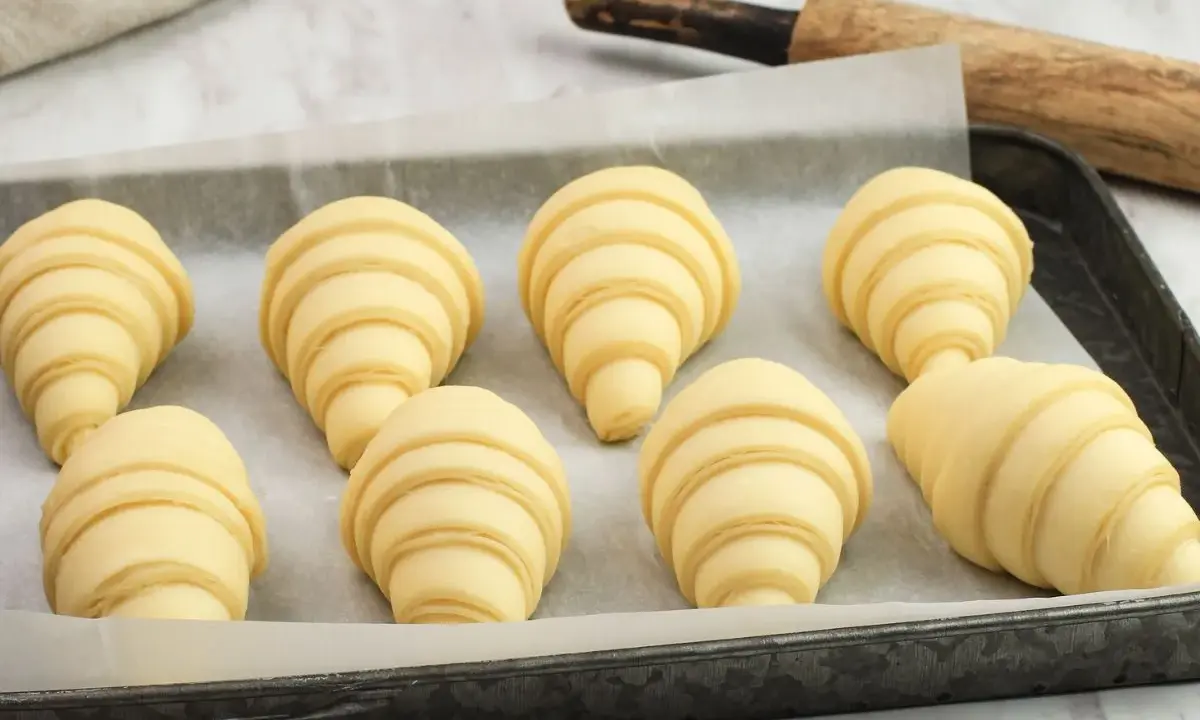 Ocho croissants caseros listos para hornear en una bandeja. Se ven deliciosos y prometen un gran desayuno.