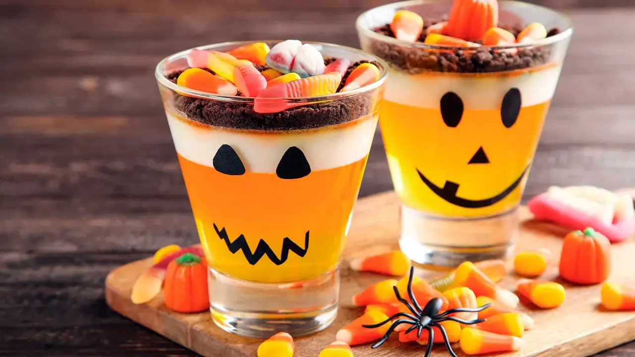 Deliciosa gelatina Halloween para niños, con capas de naranja y blanco, decorada como calabaza y cubierta de dulces.
