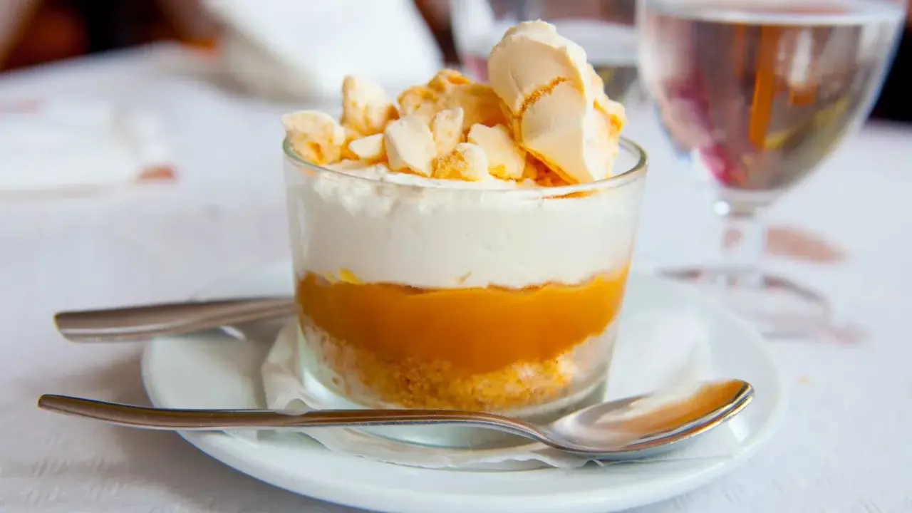 Postre en vaso con capas de galleta, crema y mermelada de mango, coronado con merengue crujiente, como un polvito canario.