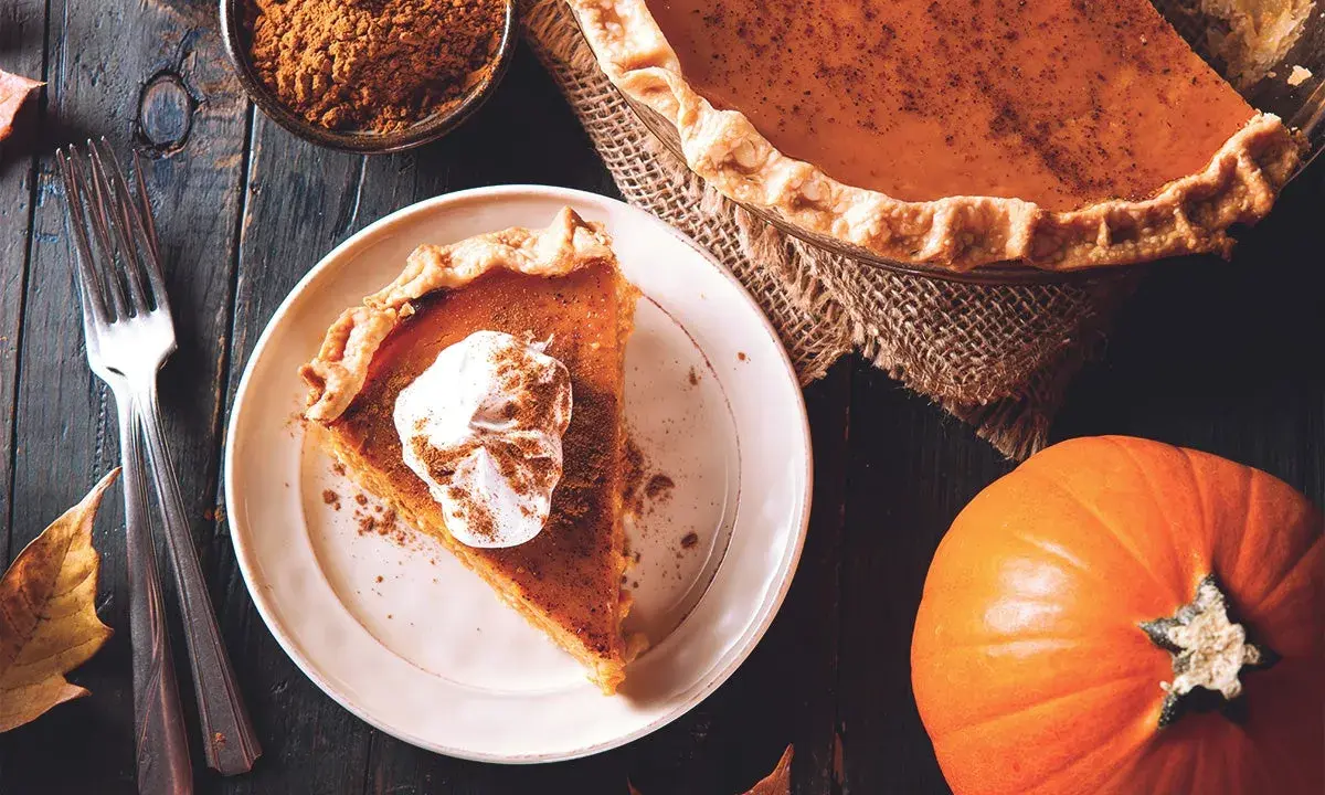 Un trozo de pastel de calabaza con crema batida y canela, junto a una calabaza entera y especias. ¡Deliciosos postres con calabaza!