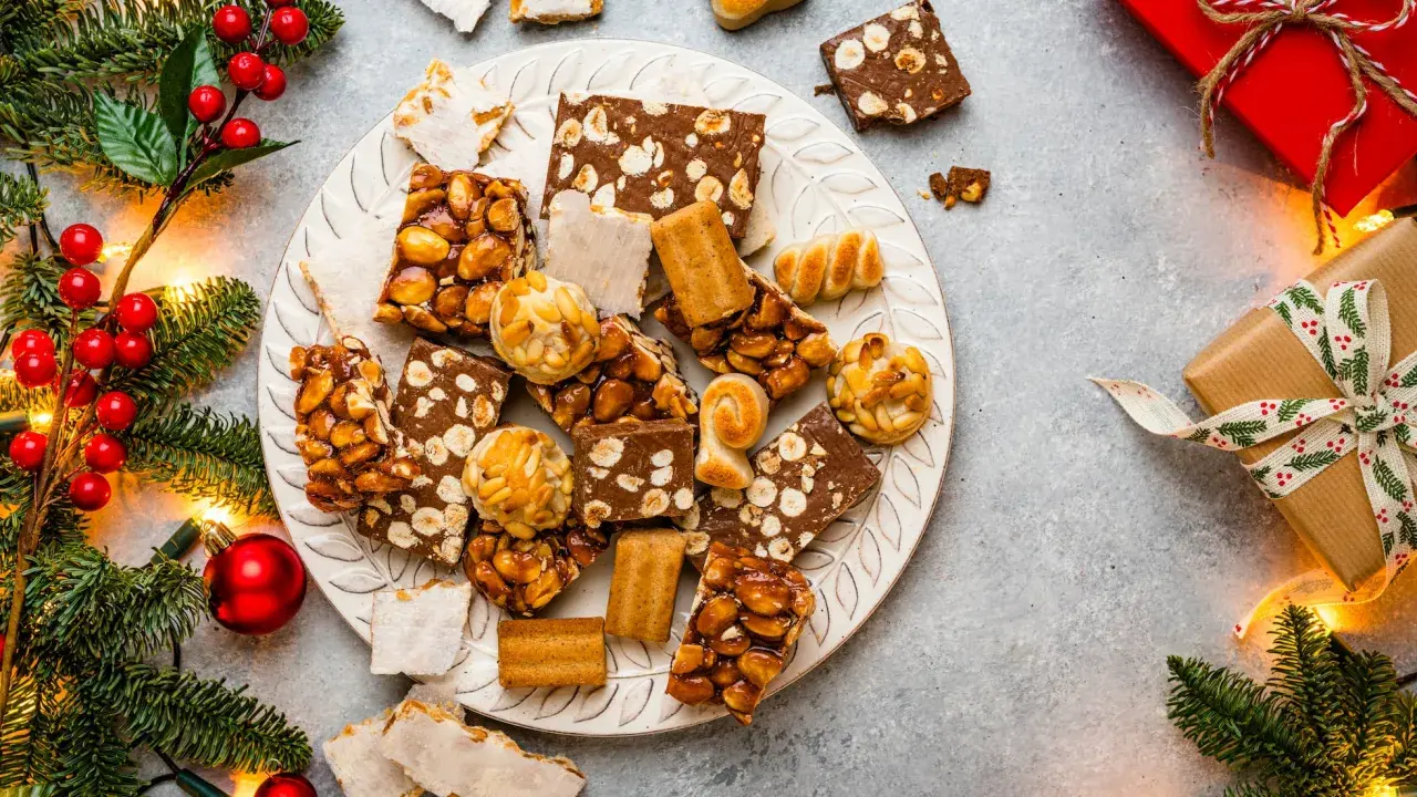Plato con variedad de postres navideños, turrones, mazapanes y dulces de frutos secos, rodeado de ramas de abeto y luces.