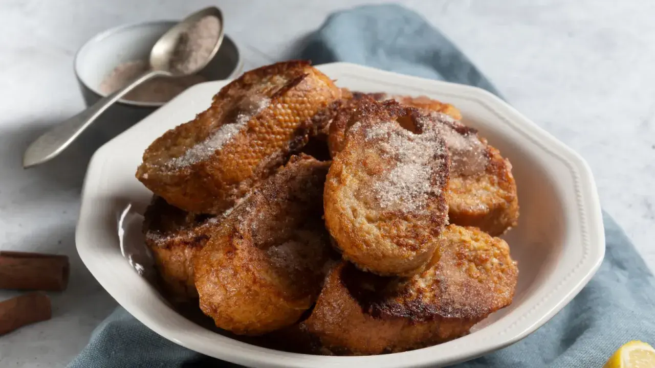 Un plato blanco lleno de torrijas españolas doradas y espolvoreadas con azúcar y canela.