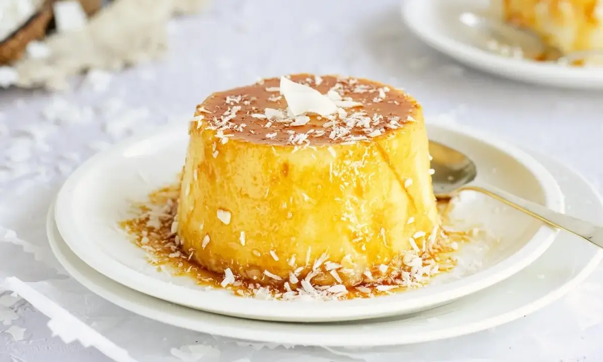 Delicioso flan de coco con caramelo líquido y coco rallado, servido en un plato blanco con una cuchara dorada.