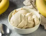 Un cremoso helado de plátano servido en un cuenco blanco, con plátanos amarillos frescos al fondo.