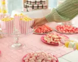 Mesa llena de dulces rosas y amarillos, palomitas en recipientes a rayas, y brochetas de malvaviscos. ¡Perfecto para aprender como hacer bolas de chuches!