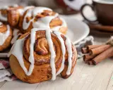 Rollo de canela cubierto de glaseado blanco, con canela en rama atada y una taza de café al fondo.