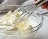 Batidor de mano mezclando crema chantilly en un bol de vidrio.