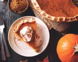 Un trozo de pastel de calabaza con crema batida y canela, junto a una calabaza entera y especias. ¡Deliciosos postres con calabaza!