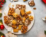 Plato con variedad de postres navideños, turrones, mazapanes y dulces de frutos secos, rodeado de ramas de abeto y luces.