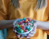 Manos sostienen un cuenco lleno de chuches saludables para niños: gomitas de corazón, bolitas de colores y más.