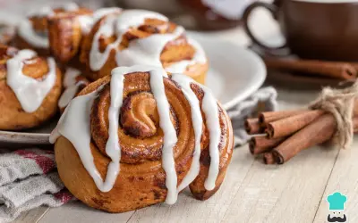 Rollo de canela cubierto de glaseado blanco, con canela en rama atada y una taza de café al fondo.