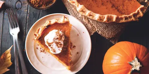 Un trozo de pastel de calabaza con crema batida y canela, junto a una calabaza entera y especias. ¡Deliciosos postres con calabaza!
