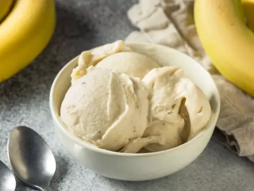 Un cremoso helado de plátano servido en un cuenco blanco, con plátanos amarillos frescos al fondo.
