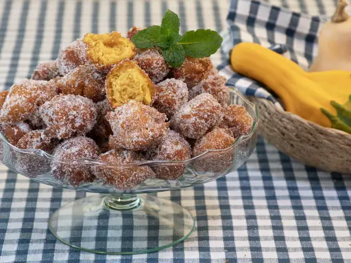Un tazón lleno de buñuelos de calabaza cubiertos de azúcar, con una ramita de menta encima. Al fondo, calabazas en una cesta.