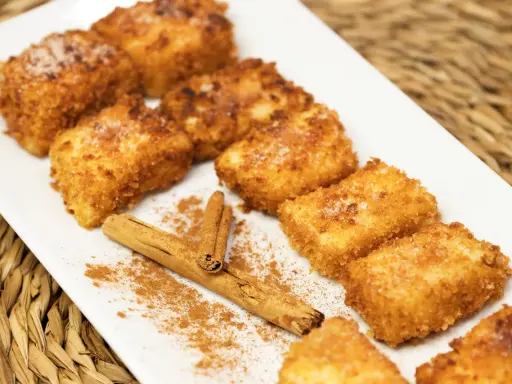 Deliciosa leche frita dorada y crujiente, espolvoreada con azúcar y canela, junto a una rama de canela.