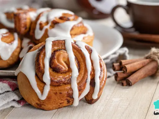 Rollo de canela cubierto de glaseado blanco, con canela en rama atada y una taza de café al fondo.