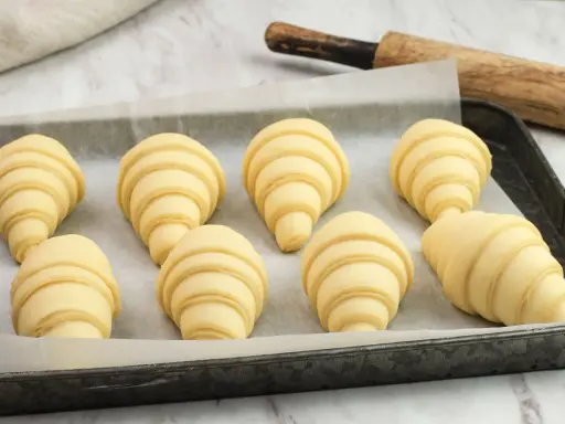 Ocho croissants caseros listos para hornear en una bandeja. Se ven deliciosos y prometen un gran desayuno.