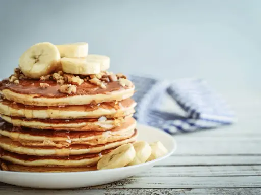 Una pila de tortitas de plátano cubiertas de miel y nueces, con rodajas de plátano fresco encima.