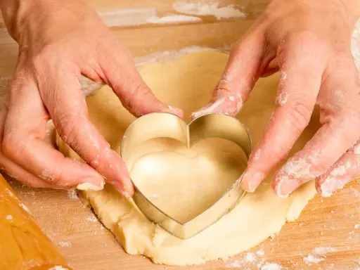 Manos cortan masa con un cortador en forma de corazón, parte de una receta de galletas caseras.