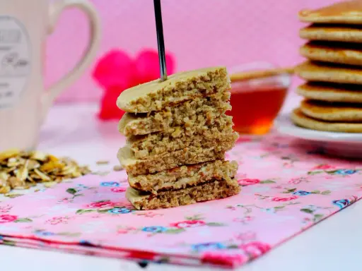 Una pila de tortitas de avena, con un tenedor atravesado, sobre una servilleta floral rosa. Al fondo, una taza y miel.