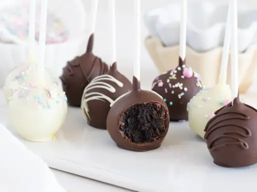 Deliciosos cake pops cubiertos de chocolate, algunos con chispas de colores y otros con glaseado blanco. Uno muestra su interior esponjoso.