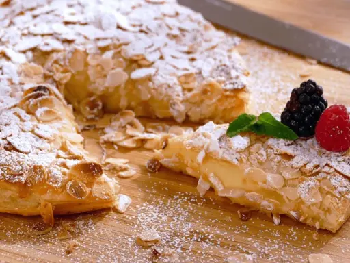 Deliciosos postres con hojaldre, cubiertos de almendras laminadas y azúcar glas. Una porción muestra un relleno cremoso y frutos rojos.
