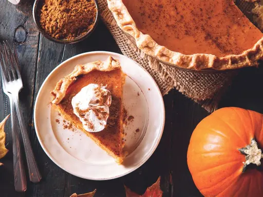 Un trozo de pastel de calabaza con crema batida y canela, junto a una calabaza entera y especias. ¡Deliciosos postres con calabaza!