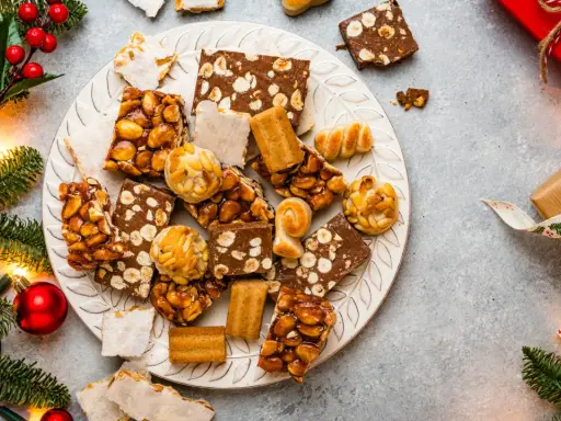 Plato con variedad de postres navideños, turrones, mazapanes y dulces de frutos secos, rodeado de ramas de abeto y luces.
