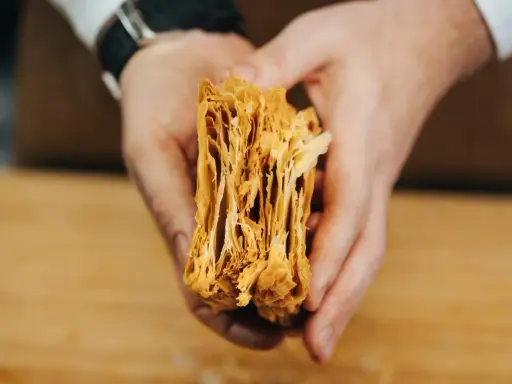 Manos sostienen un trozo de hojaldre casero, mostrando sus finas y crujientes capas doradas.