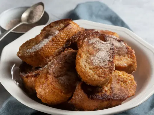 Un plato blanco lleno de torrijas españolas doradas y espolvoreadas con azúcar y canela.