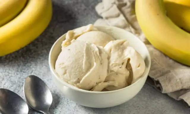 Un cremoso helado de plátano servido en un cuenco blanco, con plátanos amarillos frescos al fondo.
