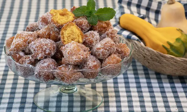 Un tazón lleno de buñuelos de calabaza cubiertos de azúcar, con una ramita de menta encima. Al fondo, calabazas en una cesta.