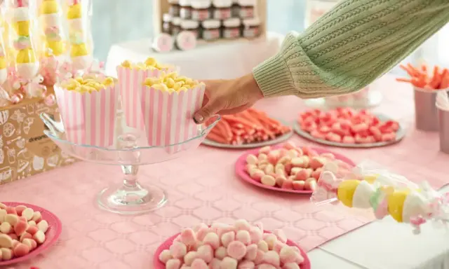 Mesa llena de dulces rosas y amarillos, palomitas en recipientes a rayas, y brochetas de malvaviscos. ¡Perfecto para aprender como hacer bolas de chuches!