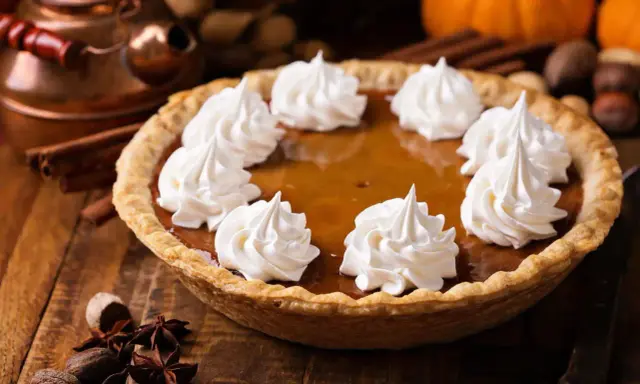 Pastel de calabaza decorado con crema batida, listo para los postres de Acción de Gracias. Especias y calabazas al fondo.