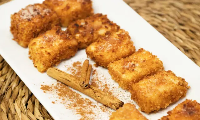 Deliciosa leche frita dorada y crujiente, espolvoreada con azúcar y canela, junto a una rama de canela.