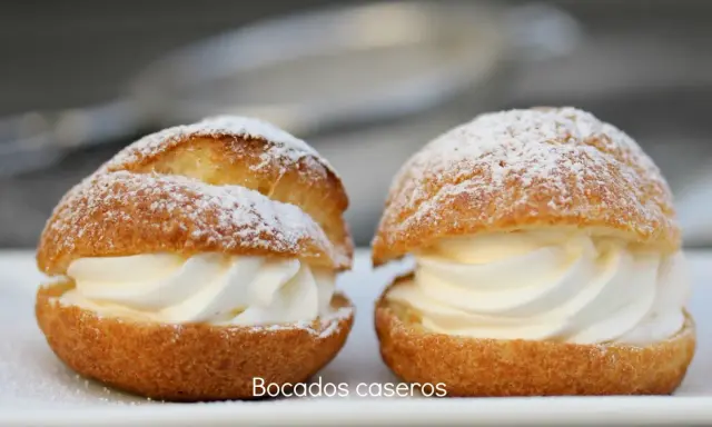 Dos bocados caseros de pasta choux, rellenos de crema batida y espolvoreados con azúcar glas.