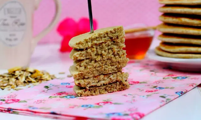 Una pila de tortitas de avena, con un tenedor atravesado, sobre una servilleta floral rosa. Al fondo, una taza y miel.