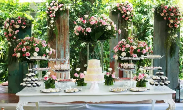 Mesa de dulces con pastel de tres pisos, cupcakes y postres variados, decorada con abundantes flores rosas y blancas. Ideal para hacer flores de golosinas.