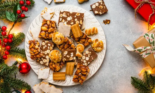 Plato con variedad de postres navideños, turrones, mazapanes y dulces de frutos secos, rodeado de ramas de abeto y luces.