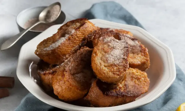 Un plato blanco lleno de torrijas españolas doradas y espolvoreadas con azúcar y canela.