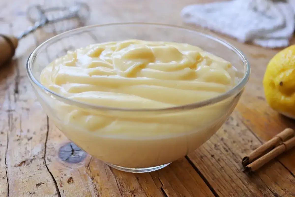 Crema pastelera cremosa en un bol de cristal, lista para rellenar postres. A su lado, un limón y canela.