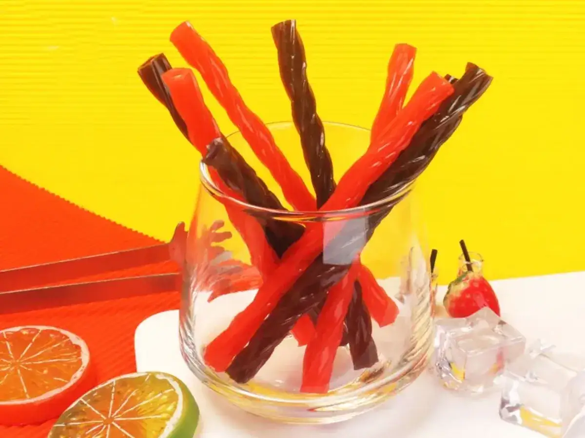Un vaso lleno de regaliz rojo y negro retorcido, como se hace el regaliz rojo. Alrededor, rodajas de naranja y lima, cubitos de hielo y una fresa.