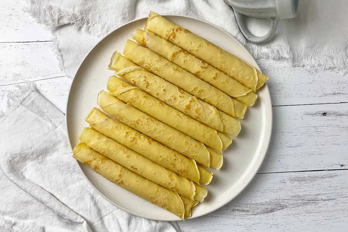 Una pila de crepes dorados enrollados en un plato blanco, listos para disfrutar. ¡Una deliciosa receta de crepes!