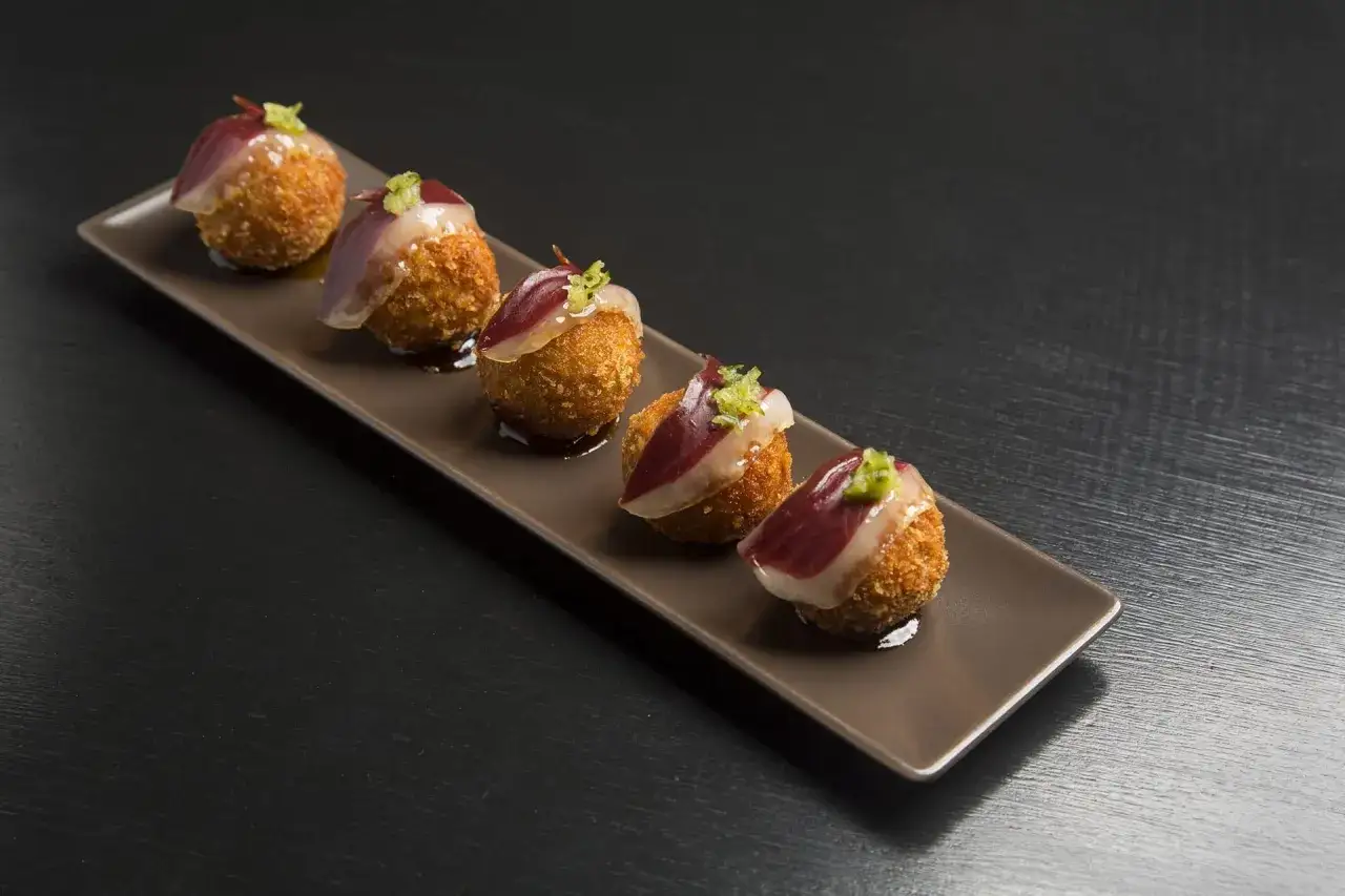 Cinco croquetas de jamón, receta española, doradas y crujientes, coronadas con jamón y un toque verde.