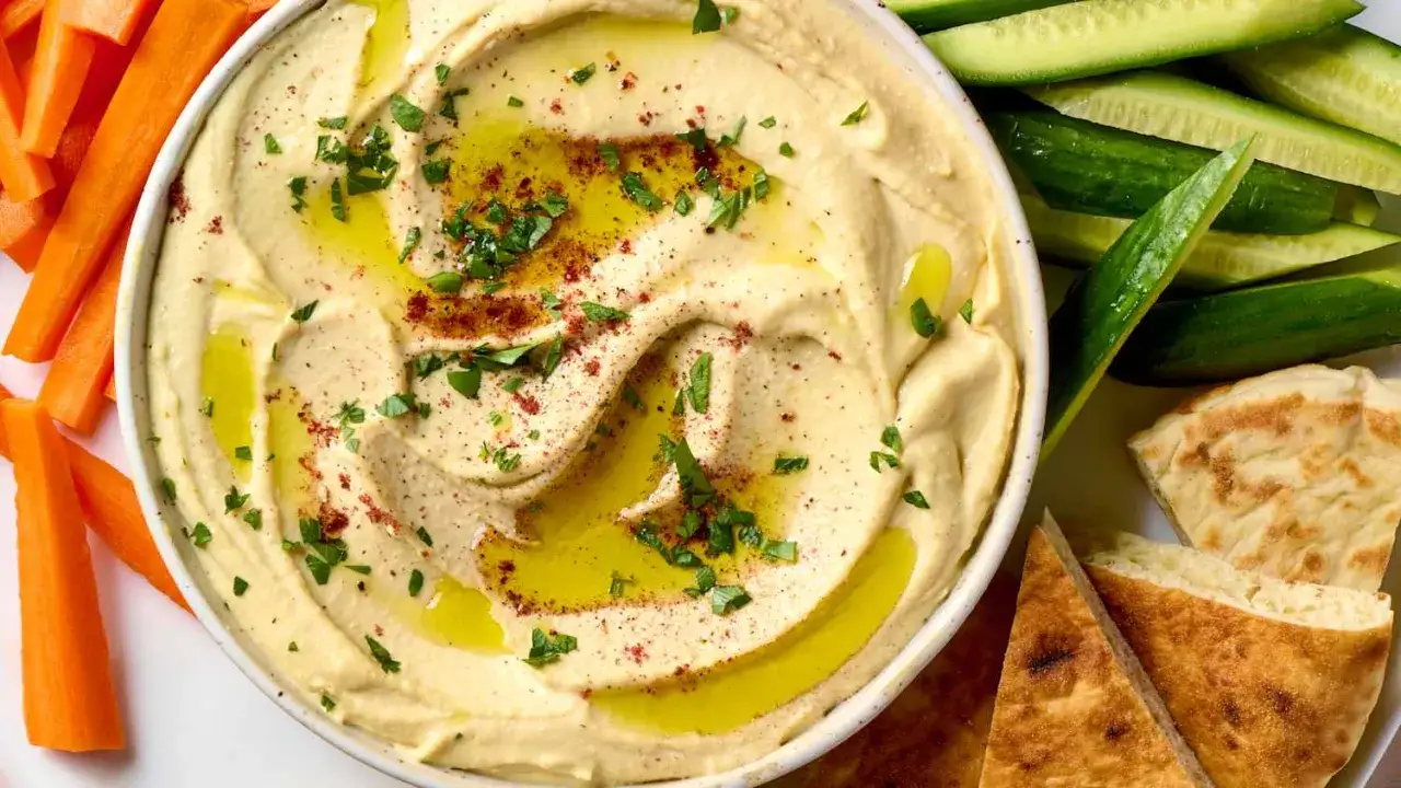 Hummus cremoso con aceite de oliva, pimentón y perejil, acompañado de bastones de zanahoria, pepino y pan pita. ¡Perfecto para aprender como hacer hummus!