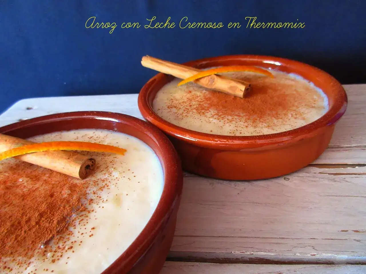 Dos cuencos de barro con cremoso arroz con leche Thermomix, espolvoreados con canela y decorados con canela en rama y piel de naranja.