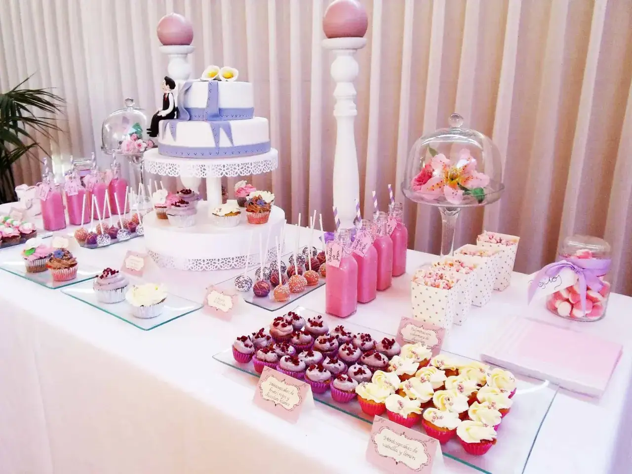 Mesa dulce con tarta de boda, cupcakes, cake pops, botellas de batido de fresa y tarros de tarta de gominolas.